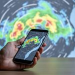 “Traveler holding a smartphone showing a non-labeled radar visualization with green, yellow, and red precipitation zones, suggesting real-time storm movement toward Quebec.”